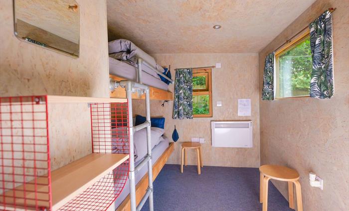 inside a wooden cabin with bunk beds, a ladder made of scaffolding, two windows with curtains and on the walls there is a small shelf, a mirror and a